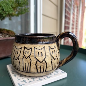 May include: A handmade ceramic mug with a brown rim and handle, featuring a repeating cat design etched into the tan surface. The mug sits on a white square coaster with a blue pattern. The mug is approximately 4 inches tall.