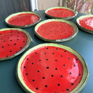 May include: A set of six ceramic plates with a watermelon design. Each plate features a bright red center with black seed details and a green rim. The plates are arranged on a dark green surface.