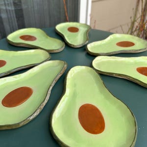 May include: A collection of ceramic avocado-shaped plates. Each plate features a light green exterior with a brown seed in the center. The plates are arranged on a dark green surface, showcasing their unique design and color palette.