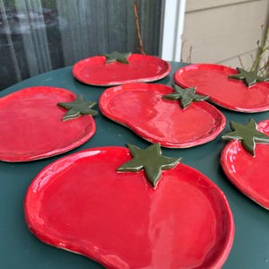 May include: A collection of red ceramic plates shaped like tomatoes, each with a green star-shaped stem. The plates are arranged on a green surface, showcasing their glossy finish and unique design. These plates are ideal for serving appetizers or small dishes.