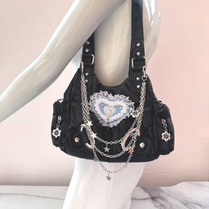 May include: Black handbag with silver chain and star accents. The bag features a heart-shaped patch with blue and white details. Silver hardware and flower charms adorn the sides. The bag is carried by a mannequin.