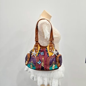 Puede incluir: Un bolso de hombro multicolor con paneles en marrón, amarillo, morado y verde azulado. El bolso está decorado con dijes plateados en forma de estrella, apliques de mariposas y pequeños remaches circulares. Incluye correas marrones y cremalleras metálicas.