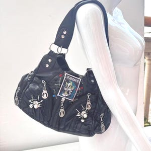 Pode incluir: Bolsa de ombro em couro preto com detalhes de aranha e pingentes prateados. A bolsa apresenta um patch decorativo com texto japonês e é adornada com alfinetes de segurança. Possui uma alça de ombro larga e ferragens prateadas.