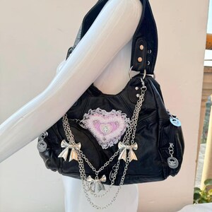 May include: Black shoulder bag with silver bow and chain accents. The bag features a heart-shaped patch with a cross design and a decorative trim. The bag has a top handle and a zipper pocket.