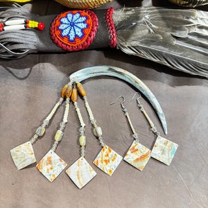 May include: A curved iridescent shell hair accessory paired with a set of matching earrings. The earrings feature diamond-shaped shell pendants suspended from cream-colored seed beads, clear crystals, and brown wooden beads on silver-toned hooks.