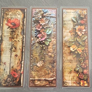 May include: Three vintage-style bookmarks with floral designs. The bookmarks are printed on paper with a textured background. The first bookmark features a red poppy flower with a compass design. The second bookmark features a variety of flowers in shades of pink, purple, and white. The third bookmark features a variety of flowers in shades of orange, yellow, and brown.
