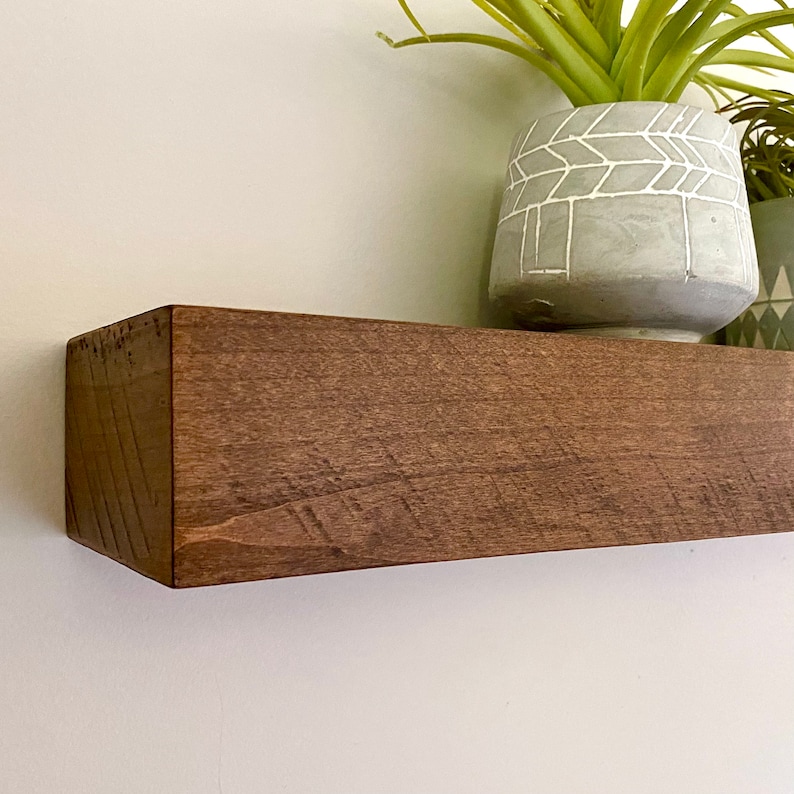 May include: A dark brown wooden shelf with a slightly rough texture. The shelf is mounted on a white wall and has a plant in a white planter with a geometric pattern on it.