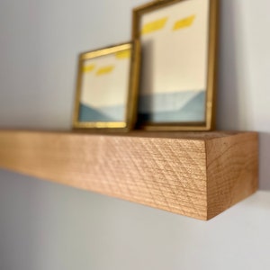 May include: A wooden shelf with a natural finish, featuring two gold picture frames with abstract artwork. The shelf is mounted on a white wall.