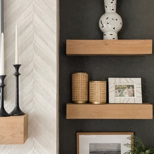 May include: Two wooden shelves with a white vase, two woven candle holders, and a framed photo of a wedding ceremony. The shelves are mounted on a black wall with white and gray herringbone tile.