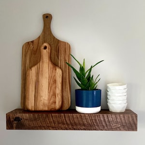 May include: A wooden shelf with a dark brown stain, holding two wooden cutting boards and a potted succulent plant with blue and white planter. The plant is a small aloe vera with green leaves. There are also four white ceramic bowls stacked on top of each other.