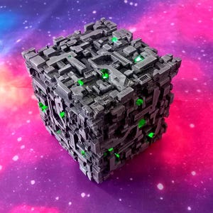 May include: A detailed, gray cube with a complex, textured surface and embedded green lights. The cube has a futuristic, industrial design, set against a vibrant purple and pink space-themed background.
