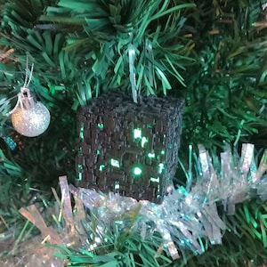 May include: A black cube-shaped Christmas ornament with a green light inside, hanging on a green artificial Christmas tree.