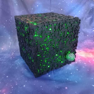 May include: A black, cube-shaped model with intricate, geometric detailing and glowing green accents. A spherical, textured element is attached to one side. The model is set against a starry, space-themed backdrop.