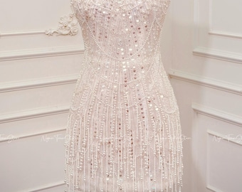 May include: A strapless, white beaded mini dress on a mannequin, adorned with sequins and dangling bead fringe. A gold crown sits atop the mannequin's head. The dress is displayed against a white wall.