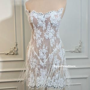 Könnte beinhalten: Ein trägerloses Mini-Kleid aus weißer Spitze mit floralen Mustern und Fransen. Das Kleid wird auf einer Schaufensterpuppe präsentiert, die sein aufwendiges Design und seine Textur hervorhebt. Das Kleid hat einen wellenförmigen Ausschnitt und ein transparentes Unterkleid.