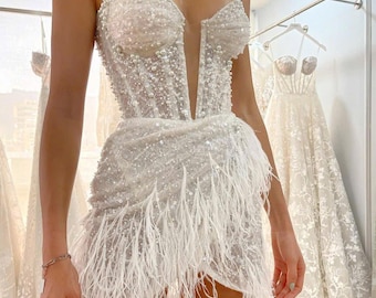 May include: A white, embellished mini dress with a sweetheart neckline and pearl-like accents. The bodice is covered in sequins and beads, and the skirt features a feathered hemline. The dress has thin, beaded straps.