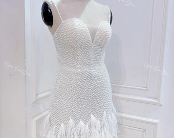 May include: A white sleeveless dress with a beaded bodice and a feather trim. The dress has a sweetheart neckline and a fitted silhouette.