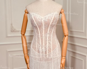 May include: A cream-colored, strapless dress with intricate lace detailing and a fringe hemline. The dress is displayed on a wooden mannequin. The bodice features vertical lines and sparkling embellishments. The dress is a formal wear item.