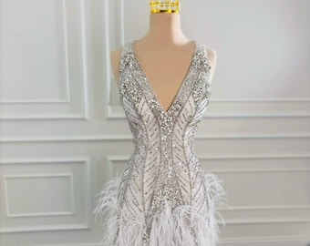 May include: A glamorous, short, white dress with a V-neck, heavily embellished with silver sequins and beads. The dress features a feathered skirt and is displayed on a mannequin with a gold crown. The dress is designed for special occasions.