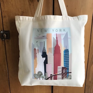 May include: A white canvas tote bag with a colorful illustration of the New York City skyline, including the Statue of Liberty and the Brooklyn Bridge. The text "NEW YORK" is printed in black at the top of the illustration.