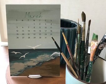 May include: A March calendar card featuring a painted coastal scene with seagulls flying over a sandy beach and ocean waves. The calendar is displayed on a white stand next to a light blue mug filled with various paintbrushes and a dark blue vase.
