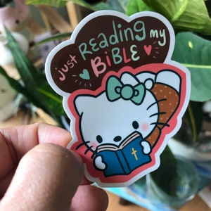 Könnte beinhalten: Aufkleber mit einer Comic-Katze, die eine blaue Bibel mit einem goldenen Kreuz liest. Der Aufkleber hat den Text "just reading my BIBLE" in einem braunen, wolkenförmigen Design mit Herzen und einer Schleife.