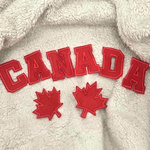 Peut inclure: Gros plan sur un tissu gris clair et texturé avec le mot "CANADA" en lettres rouges. Deux écussons de feuilles d'érable rouges sont en dessous. Le tissu a un aspect doux et moelleux.
