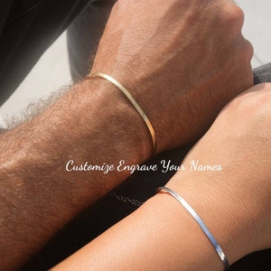 May include: Two simple gold and silver bracelets on wrists. The bracelets are thin and flat. The text "Customize Engrave Your Names" is visible on the image.