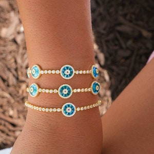 May include: Three gold bracelets with blue and white evil eye charms. Each bracelet is adorned with small clear stones.