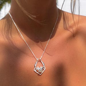 May include: A silver necklace with a pendant shaped like a ring within a geometric frame. The pendant features a clear, round gemstone. The necklace is worn around the neck.