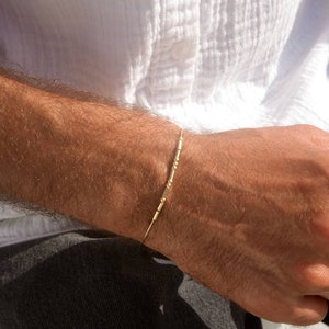 May include: A gold bracelet with a linear design featuring small, decorative segments. The bracelet is worn on a wrist, with a white shirt and dark pants in the background. The bracelet's design is simple and elegant.