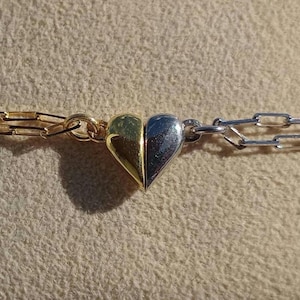 May include: A gold and silver-toned bracelet with a heart-shaped charm. The charm is split into two halves, gold and silver. The bracelet has a chain link design.