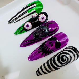 May include: Five long, pointed artificial nails with Halloween-themed designs. The nails feature a variety of colors, including green, black, and purple, with designs such as eyeballs, a skull, and a black and white spiral.