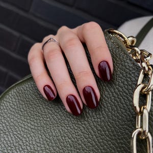 Puede incluir: Una mano con uñas ovaladas, pintadas de color burdeos intenso, descansa sobre un bolso de cuero verde oliva texturizado. Un anillo plateado está en el dedo anular. Se ve una correa de cadena dorada.