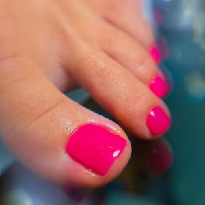 Puede incluir: Primer plano de un pie con esmalte de uñas rosa brillante en las uñas de los pies. La piel es de color melocotón claro y las uñas están cuidadosamente pintadas con un acabado brillante. El fondo está borroso, con toques de azul y blanco.