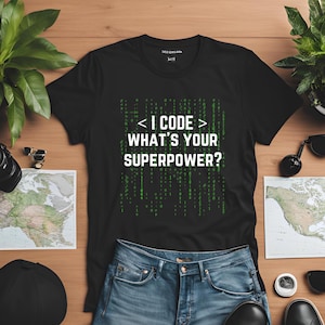 May include: Black t-shirt with the text "< I CODE > WHAT'S YOUR SUPERPOWER?" in white and green. The shirt is paired with blue jeans. The image is on a wooden surface with maps, sunglasses, and plants.