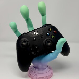May include: A black Xbox controller rests on a colorful, stylized hand-shaped stand. The stand features a gradient of blue, purple, and green, with a pink base. The controller has colored buttons and a central directional pad.
