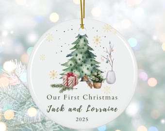 May include: A white ceramic Christmas ornament with a watercolor design. It features a green Christmas tree, a wrapped gift, and the text "Our First Christmas Jack and Lorraine 2025". The ornament hangs from a gold string.