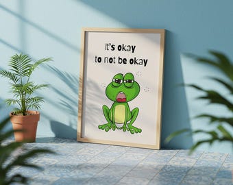 May include: A framed print with the text "It's okay to not be okay" and a cartoon frog illustration. The frog is green with a yellow belly and an open mouth. The frame is light brown.