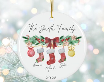 May include: A white ceramic Christmas ornament with a gold string. The ornament features the text "The Smith Family" above a festive garland with stockings, ornaments, and the year 2025.