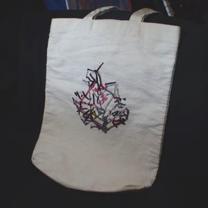 May include: Off-white tote bag with two handles. The bag features an embroidered abstract design in shades of red, black, purple, and yellow. The tote bag is made of a sturdy fabric and is suitable for carrying various items.