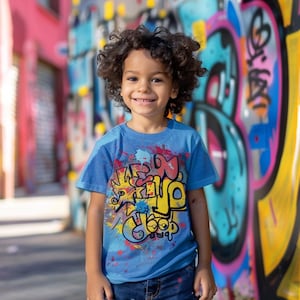 May include: A young child wearing a blue t-shirt with a colourful graffiti-style design that says "Keep it Cool". The child is standing in front of a wall with graffiti art.
