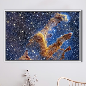 Pode incluir: Uma impressão em tela emoldurada com uma imagem vibrante dos Pilares da Criação, uma nebulosa na Nebulosa da Águia. A obra de arte mostra nuvens de poeira e gás cósmico em tons de ouro, laranja e azul, em um fundo de inúmeras estrelas.