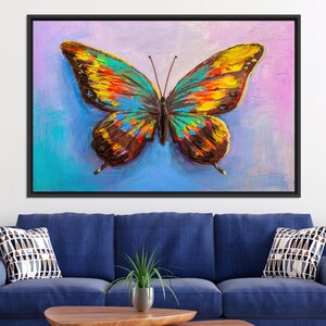 May include: A framed canvas art piece featuring a colorful butterfly. The butterfly's wings are painted with a textured, impressionistic style, showcasing shades of yellow, orange, blue, and brown against a blue and pink background.
