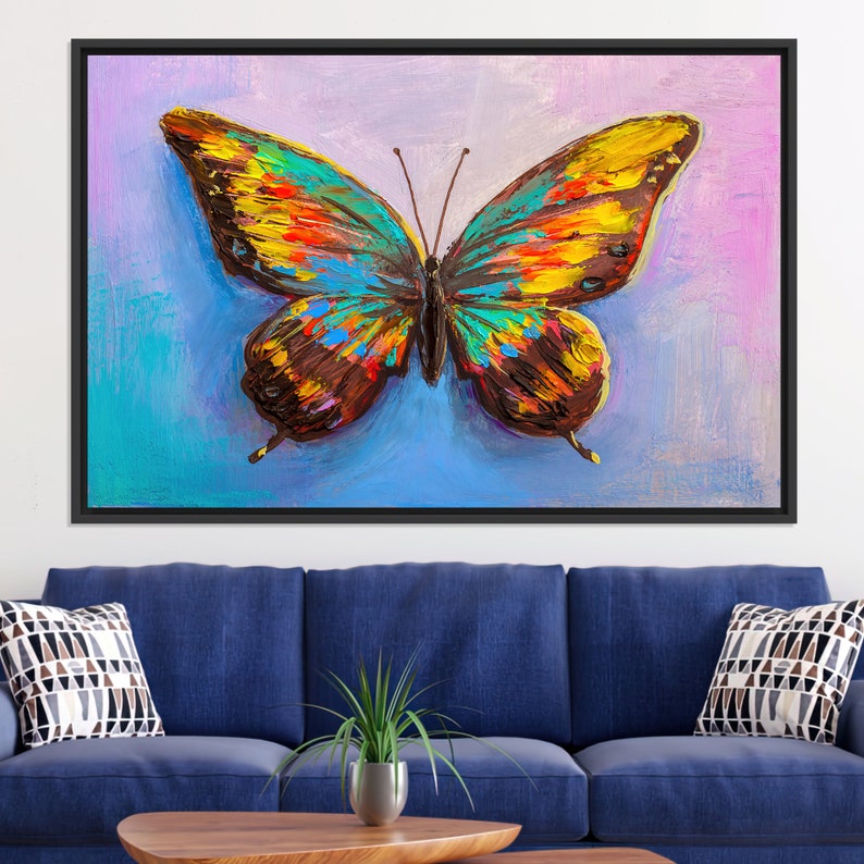 May include: A framed canvas art piece featuring a colorful butterfly. The butterfly's wings are painted with a textured, impressionistic style, showcasing shades of yellow, orange, blue, and brown against a blue and pink background.