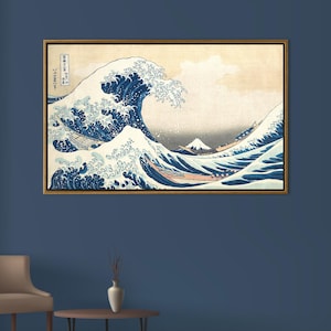 May include: Framed print of "The Great Wave off Kanagawa" by Hokusai. The artwork depicts a large ocean wave with white crests, boats, and Mount Fuji in the background. The print has a gold-colored frame and is displayed on a blue wall.