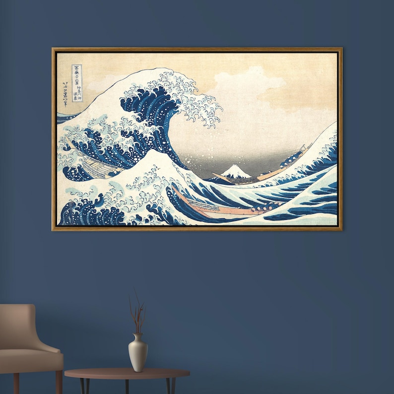 May include: Framed print of "The Great Wave off Kanagawa" by Hokusai. The artwork depicts a large ocean wave with white crests, boats, and Mount Fuji in the background. The print has a gold-colored frame and is displayed on a blue wall.