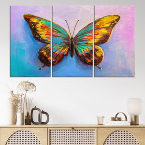 May include: A vibrant, multi-panel canvas art piece featuring a colorful butterfly. The butterfly's wings display a mix of blue, yellow, orange, and brown hues, set against a blue and purple background. The artwork is displayed above a wooden cabinet.