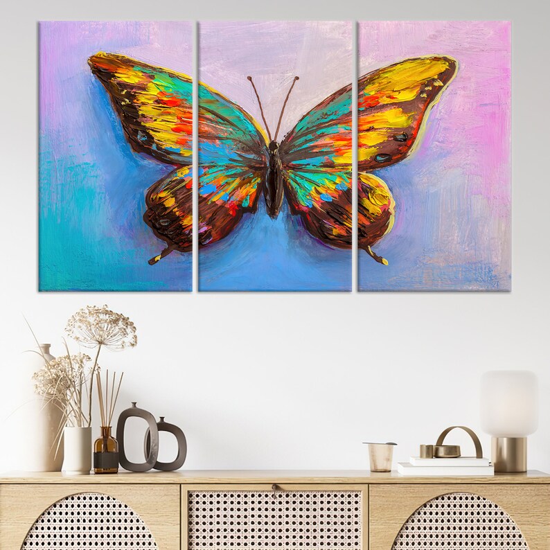 May include: A vibrant, multi-panel canvas art piece featuring a colorful butterfly. The butterfly's wings display a mix of blue, yellow, orange, and brown hues, set against a blue and purple background. The artwork is displayed above a wooden cabinet.