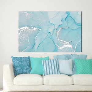 Puede incluir: Impresión artística abstracta en tonos azules y plateados, montada sobre un sofá blanco con varios cojines azules. La obra de arte presenta patrones fluidos y veteados con acentos metálicos, creando una estética moderna y serena. Los cojines añaden toques de color y textura.